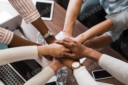 Preview: cropped view of multicultural businesspeople holding joined hands above desk with gadgets, top view