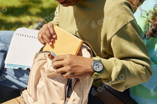 Preview: Teenage Student on Bench Putting Notebook into Backpack Outdoors