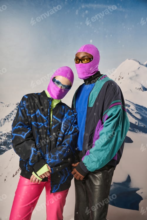 Preview: stylish young couple in warm winter outfits and balaclavas posing together on snowy backdrop