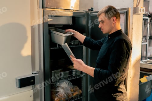 Preview: chef stands in kitchen in a black uniform and looks through what is on the list in the refrigerator