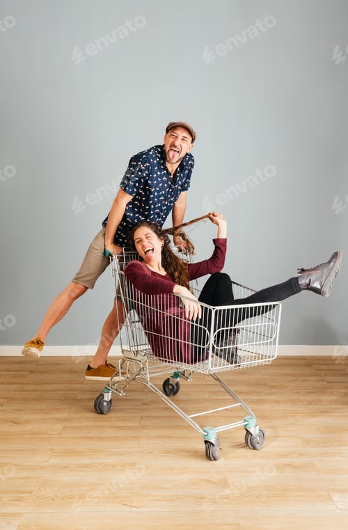 Preview: Cheerful Couple Having Fun With Shopping Cart Indoors and Sticking Their Tongues Out