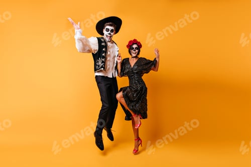 Preview: Glad zombie man in sombrero jumping on yellow background. Charming muerte girl in black dress danci