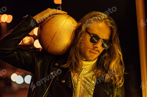 Preview: handsome young man in sunglasses and leather jacket holding golden basketball ball under yellow