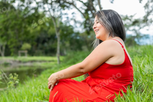 Preview: latina woman in a red dress sitting on the lawn, looking towards the horizon while smiling