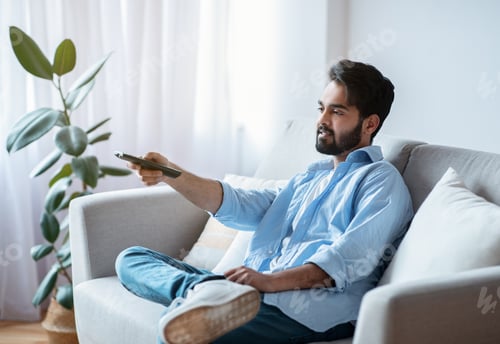 Preview: Arab man with remote controller switching channels while watching tv at home