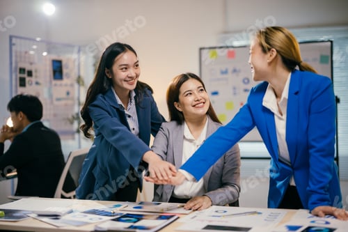 Preview: Three smiling businesswomen are putting their hands together over a desk in a modern office, showing