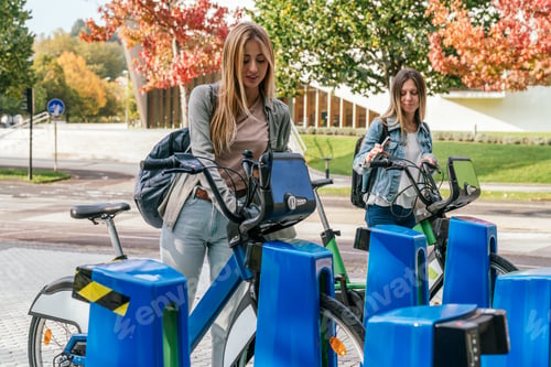 Preview: Two women taking bike for rent at bike rental station in a sunny day