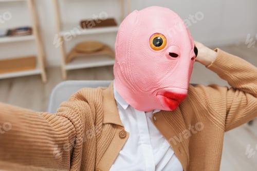 Preview: Funny blonde lady in pink fish mask warm sweater doing selfie use phone sitting in armchair at