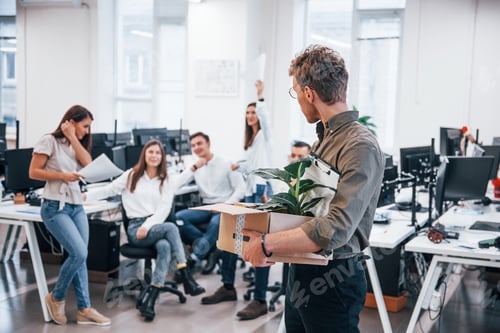 Preview: Stylish man in glasses holding box with plant in front of his colleagues in the office