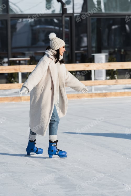 Preview: full length of joyful young woman in winter hat and coat skating on ice rink