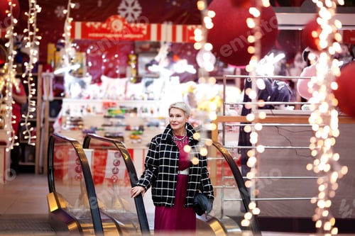 Preview: Beautiful mature woman stands on escalator in shopping center mall with New Year or Christmas decor