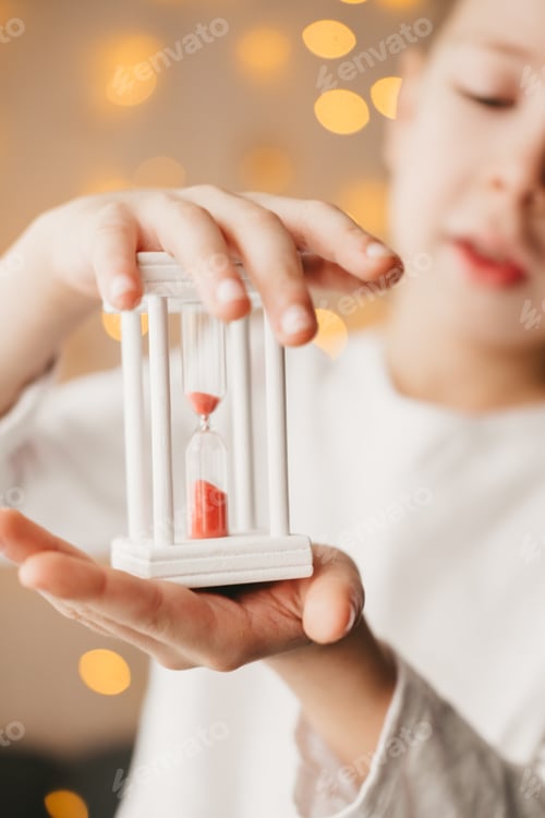 Preview: The boy is holding a white hourglass against the background of a Christmas garland.