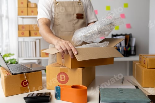 Preview: Male owner of an online store packs products into parcel boxes based on orders from online sales sit