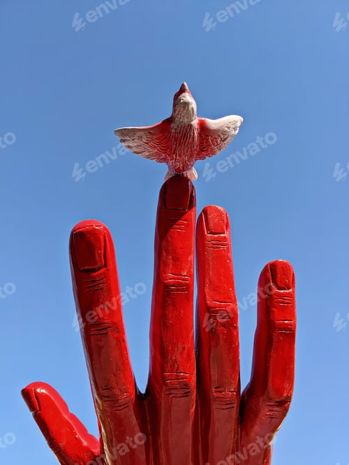Preview: Red Hand Sculpture with Bird on Finger Against Blue Sky