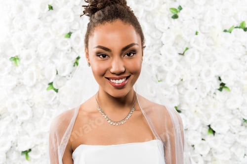 Preview: happy african american girl in white veil looking at camera near flowers