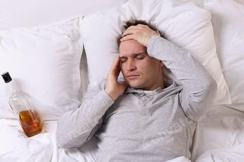 Preview: Unhappy young man with bottle of whiskey suffering from hangover on bed at home, top view