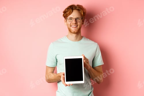 Preview: Funny redhead guy in glasses demonstrate digital tablet screen, smiling at camera, standing over