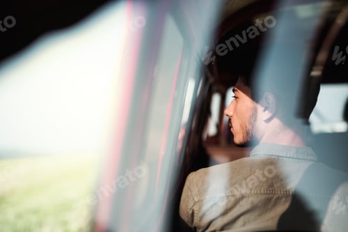 Preview: A young man sitting in a car on roadtrip through countryside, shot through glass.
