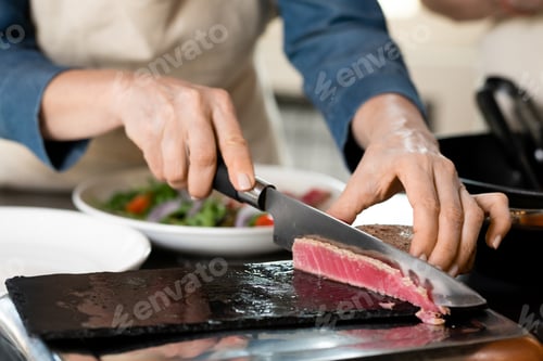 Preview: Hands of mature woman cutting piece of smoked meat on board while taking part in cooking masterclass