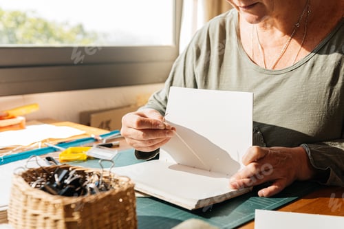 Preview: Crop of nrecognizable ederly woman sitting at desk and sewing the pages to make a handmade notebook