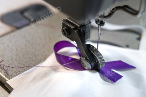 Preview: Closeup of a purple ribbon being sewn on a facemask with a sewing machine - COVID-19 awareness