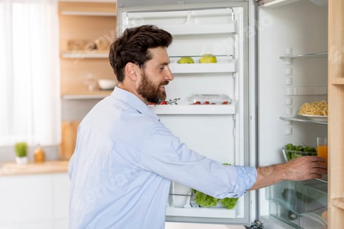 Preview: Smiling hungry handsome adult caucasian man with beard takes out food from refrigerator in white