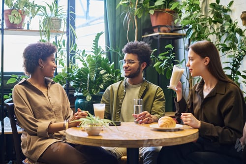 Preview: Group of intercultural zoomers having coffee and chatting by table in cozy cafe