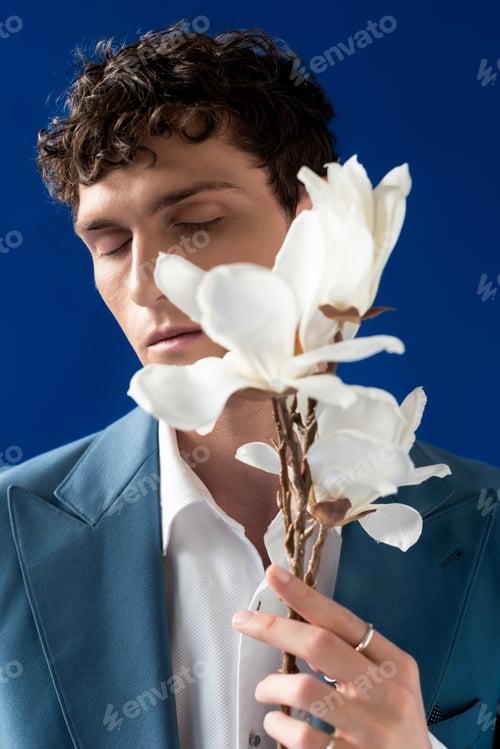 Preview: Portrait of curly young man in jacket holding blurred magnolia branch isolated on blue