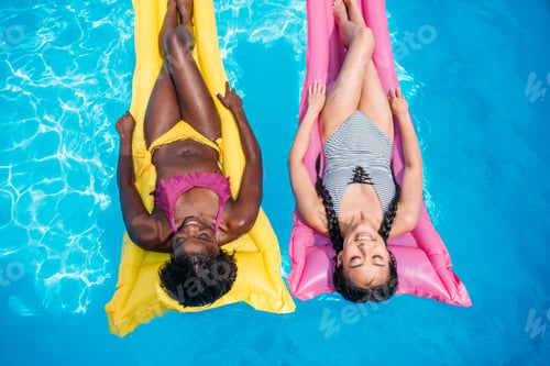 Preview: Young multiethnic women floating on inflatable mattresses in swimming pool at resort