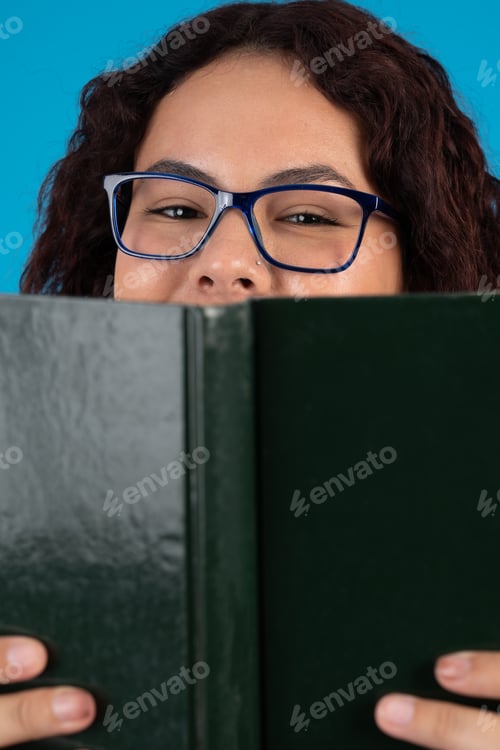 Preview: Smiling young woman with glasses, hiding half of her face behind a hardcover book