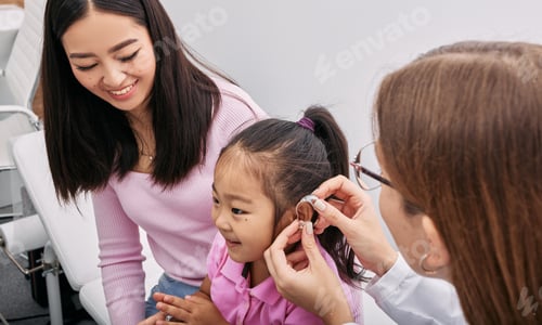 Preview: Audiologist fits hearing aid on Asian little girl's ear while visit in hearing clinic with mother.