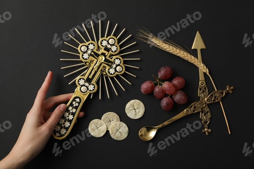 Preview: Cross in hand, liturgical bread, grape and cutlery on black background, top view
