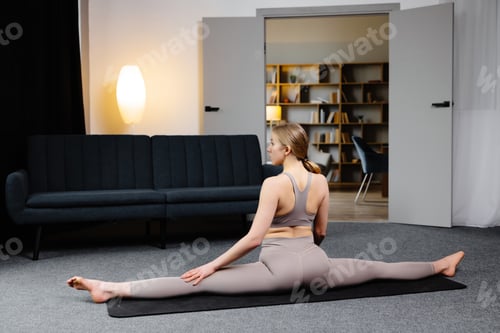 Preview: Woman in sportswear is sitting in twine on the floor in living room. Healthy lifestyle, yoga