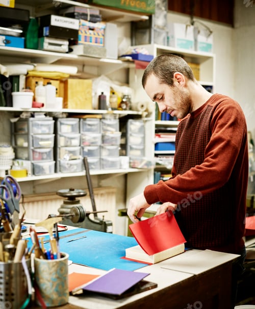 Preview: A man working in a book binding workshop, creating a red cover for freshly stitched pages.