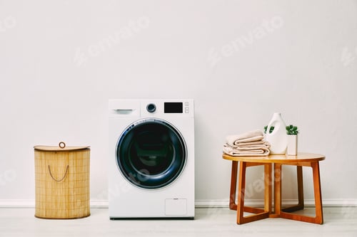 Preview: modern washing machine near laundry basket, coffee table with towels, detergent bottle and plant in