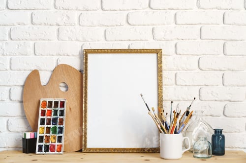 Preview: Photo frame as easel with artist's tools on wooden table against white brick wall