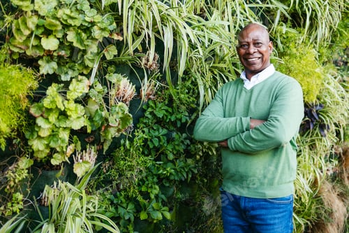 Preview: Mature man smiling with crossed arms near lush greenery