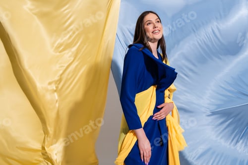 Preview: joyful young ukrainian woman in color block clothing posing near blue and yellow fabric