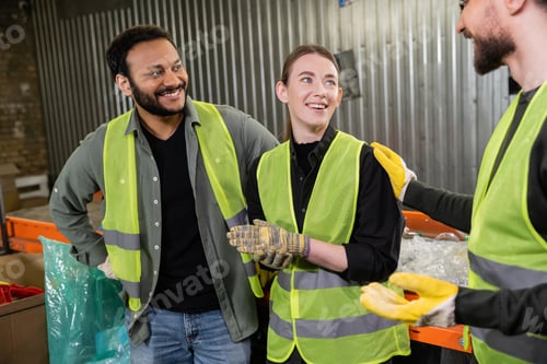 Preview: Cheerful sorter in protective vest and gloves talking to interracial colleagues while standing near