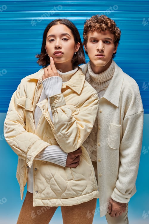 Preview: winter fashion campaign, interracial couple jackets looking at camera on blue plastic backdrop