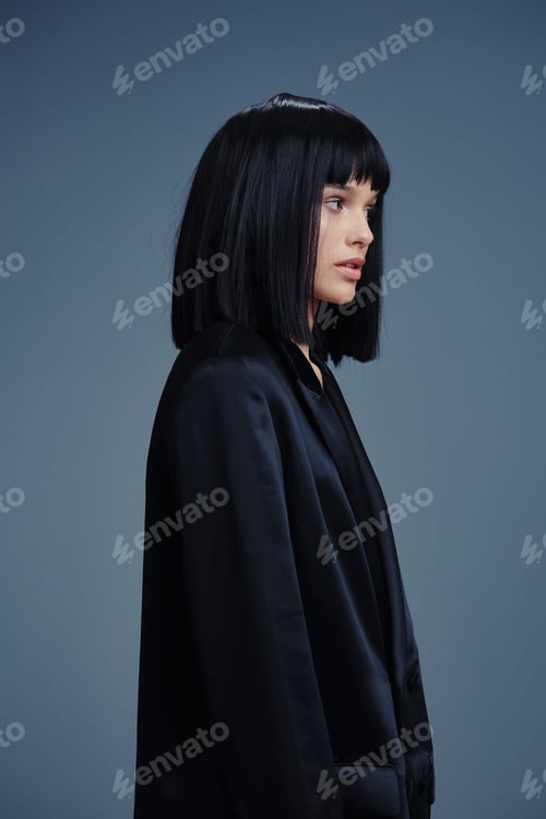 Preview: Blackhaired young woman in black jacket stands in front of gray background, looking to the side with