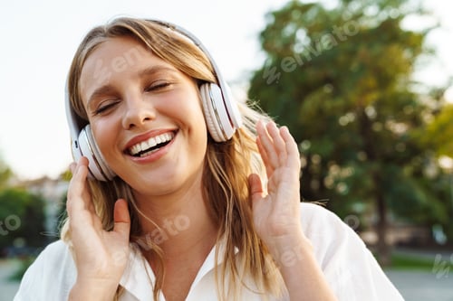 Preview: Beautiful happy woman in headphones smiling while walking in green park