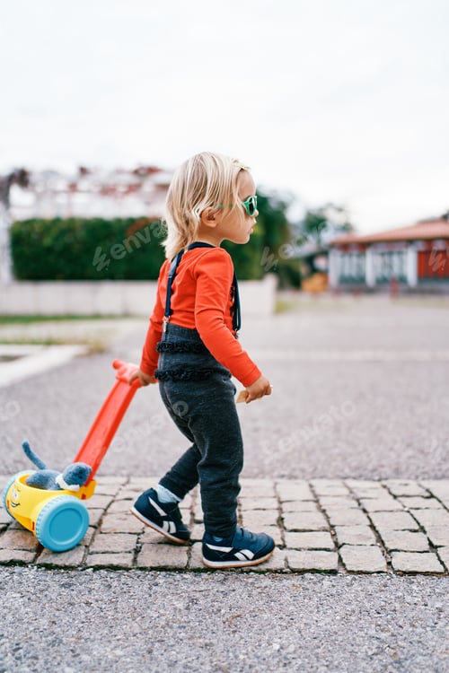Preview: Little girl is carrying a toy cart with a soft toy on the paving stones. Side view