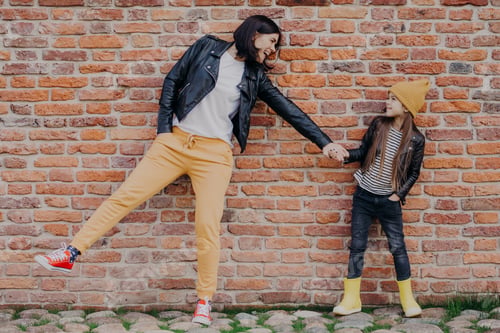 Preview: Small girl and her mother have fun together, keep hands together, pose against brick wall