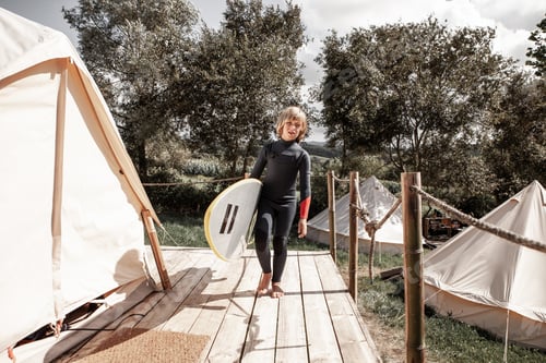 Preview: A boy dressed in a wetsuit carries his surfboard at a campsite.