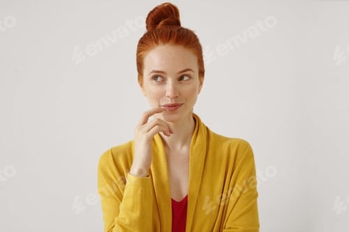 Preview: Portrait of stunning young female wearing her ginger hair in knot touching lips, looking away with s