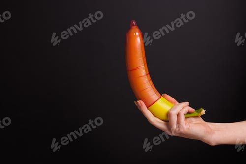 Preview: female hand holding banana with condom on dark background