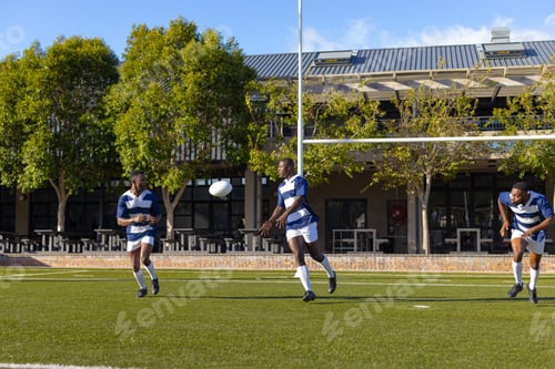 Preview: Three African American young male athletes in blue and white gear play rugby on field outdoors