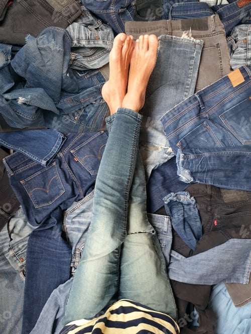 Preview: low section of woman with legs crossed at ankles sitting on pile of jeans