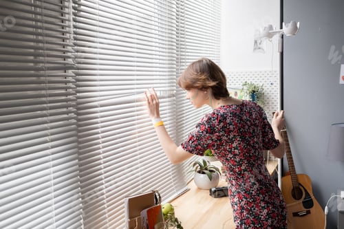 Preview: Sick young woman looking through venetian blinds
